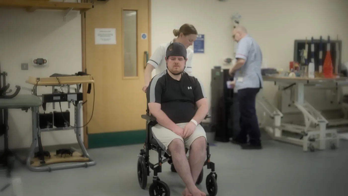 Dan, wearing a protective helmet, stands between two members of staff during a physiotherapy session