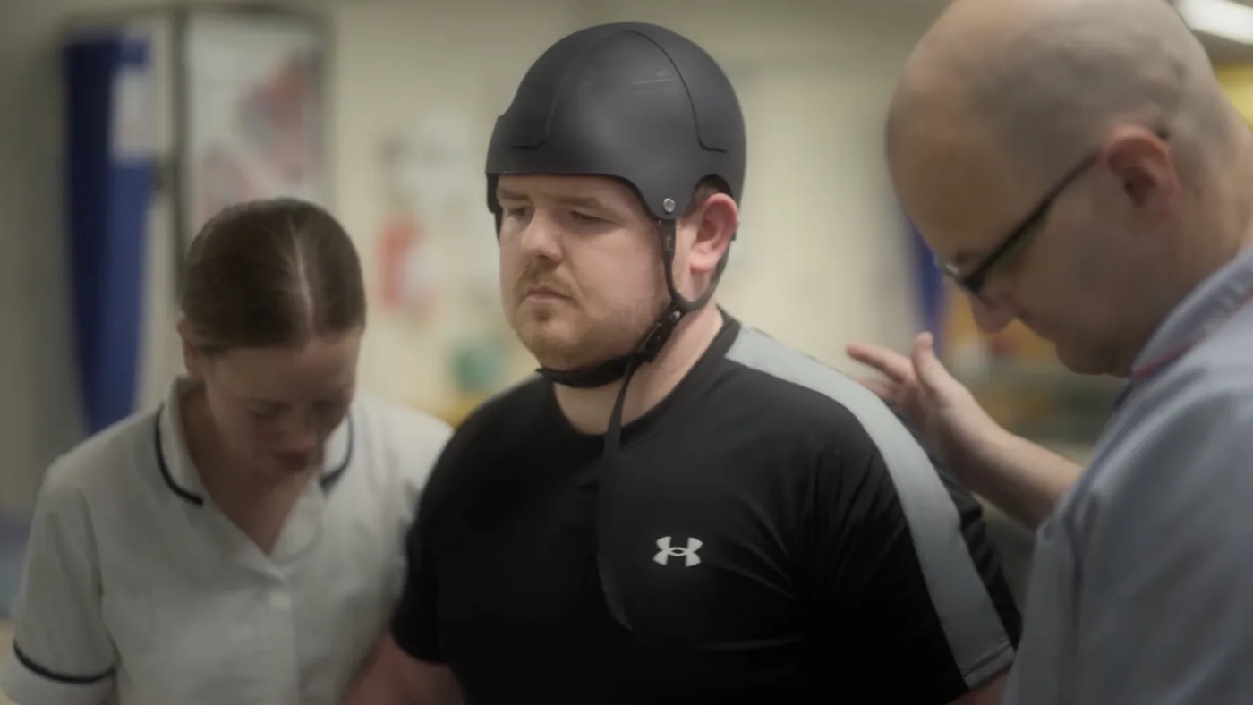 Dan sits in a wheelchair in a hospital therapy gym with a member of staff standing behind him