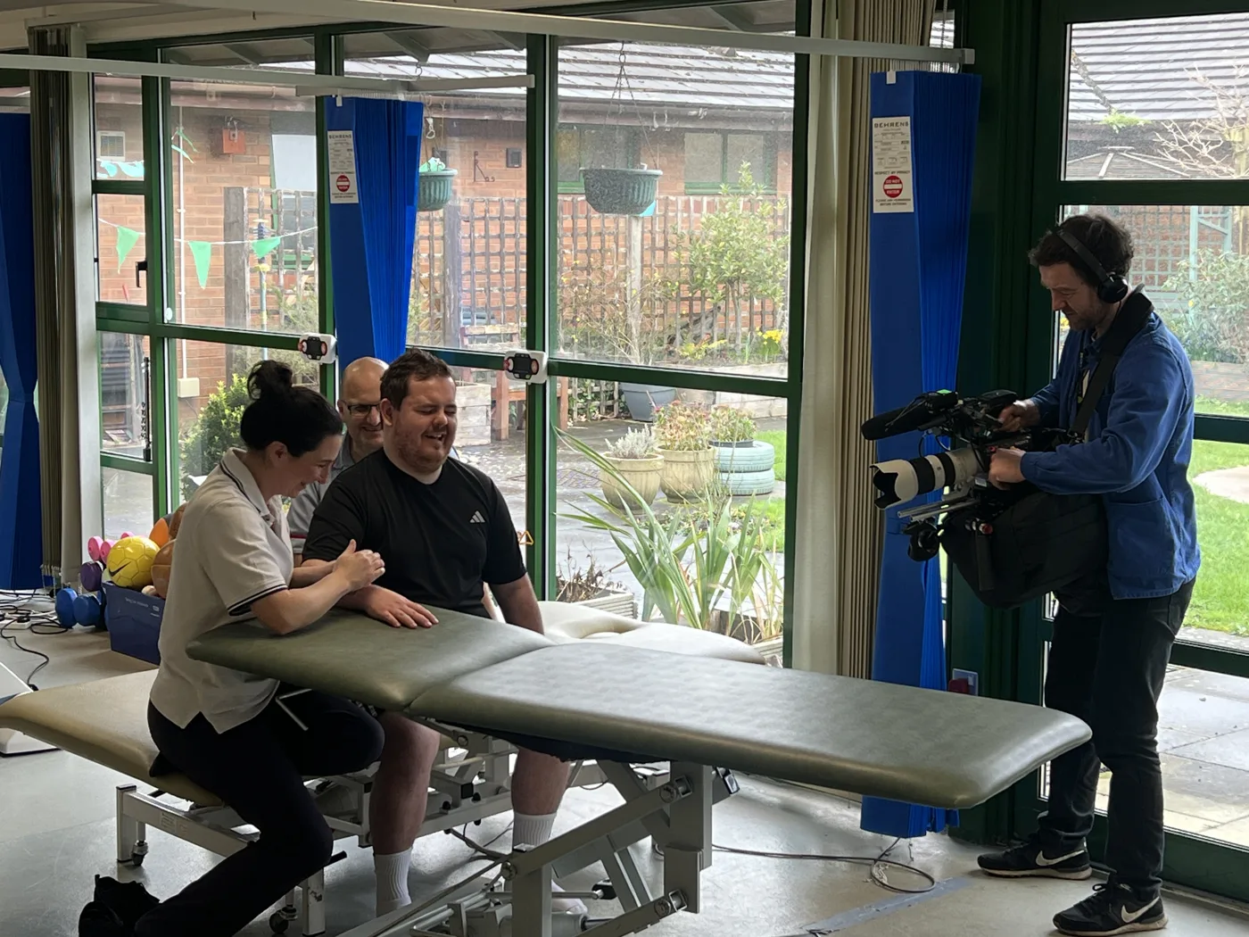 Dan sits at a treatment couch with a physiotherapist beside him while a camera operator films the session