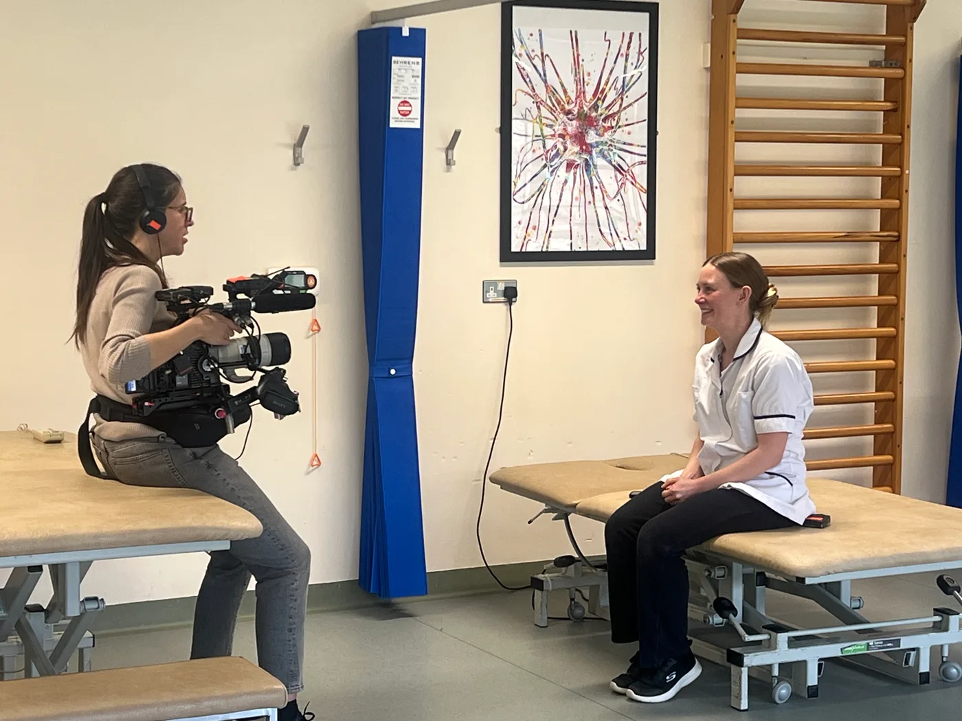 Katy Armstrong, in a white NHS tunic, sits on a treatment couch in a therapy gym while a camera operator films her