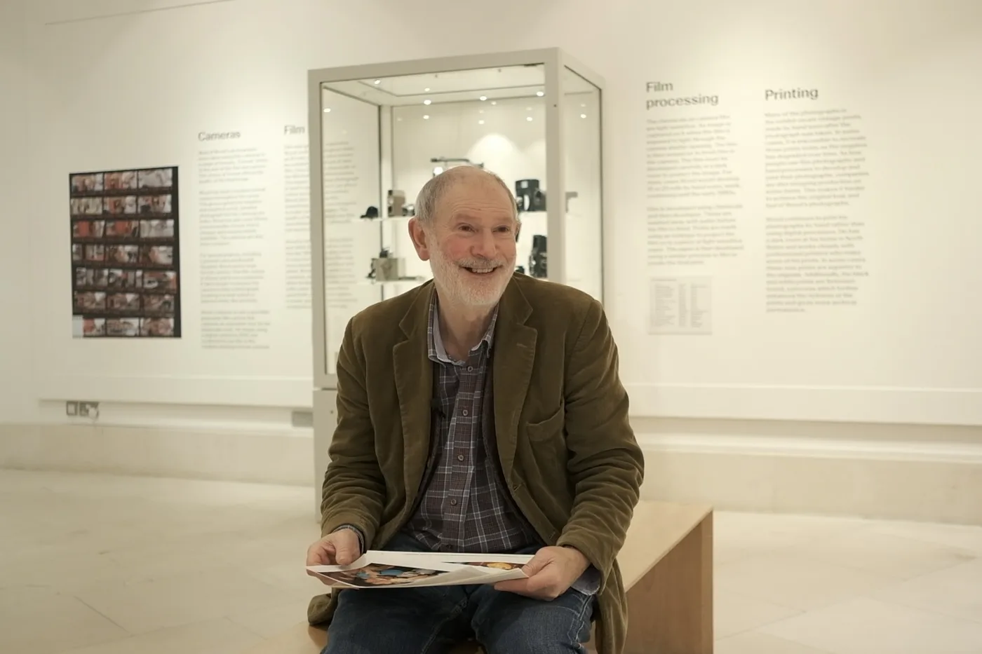 A man with a grey beard sits smiling in a gallery holding a book of photographs, with a display case of cameras behind him