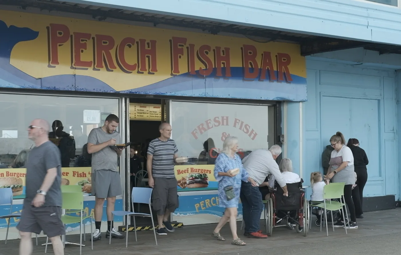 People eating outside Perch Fish Bar on the New Brighton seafront, under a hand-painted sign