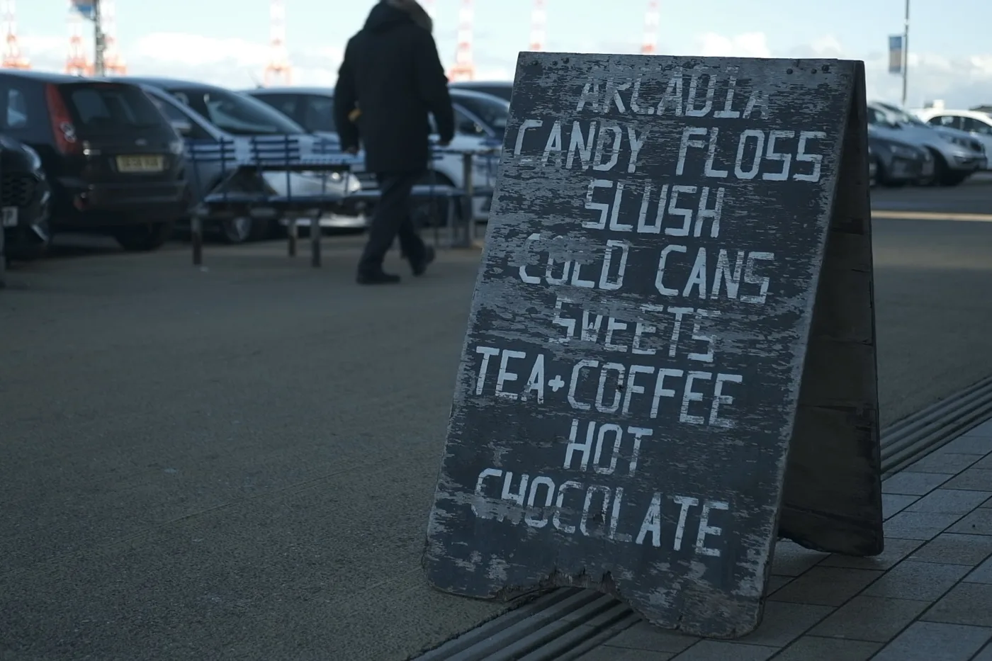 A blackboard sign outside Arcadia listing candy floss, slush, cold cans, sweets, tea and coffee and hot chocolate, with parked cars behind