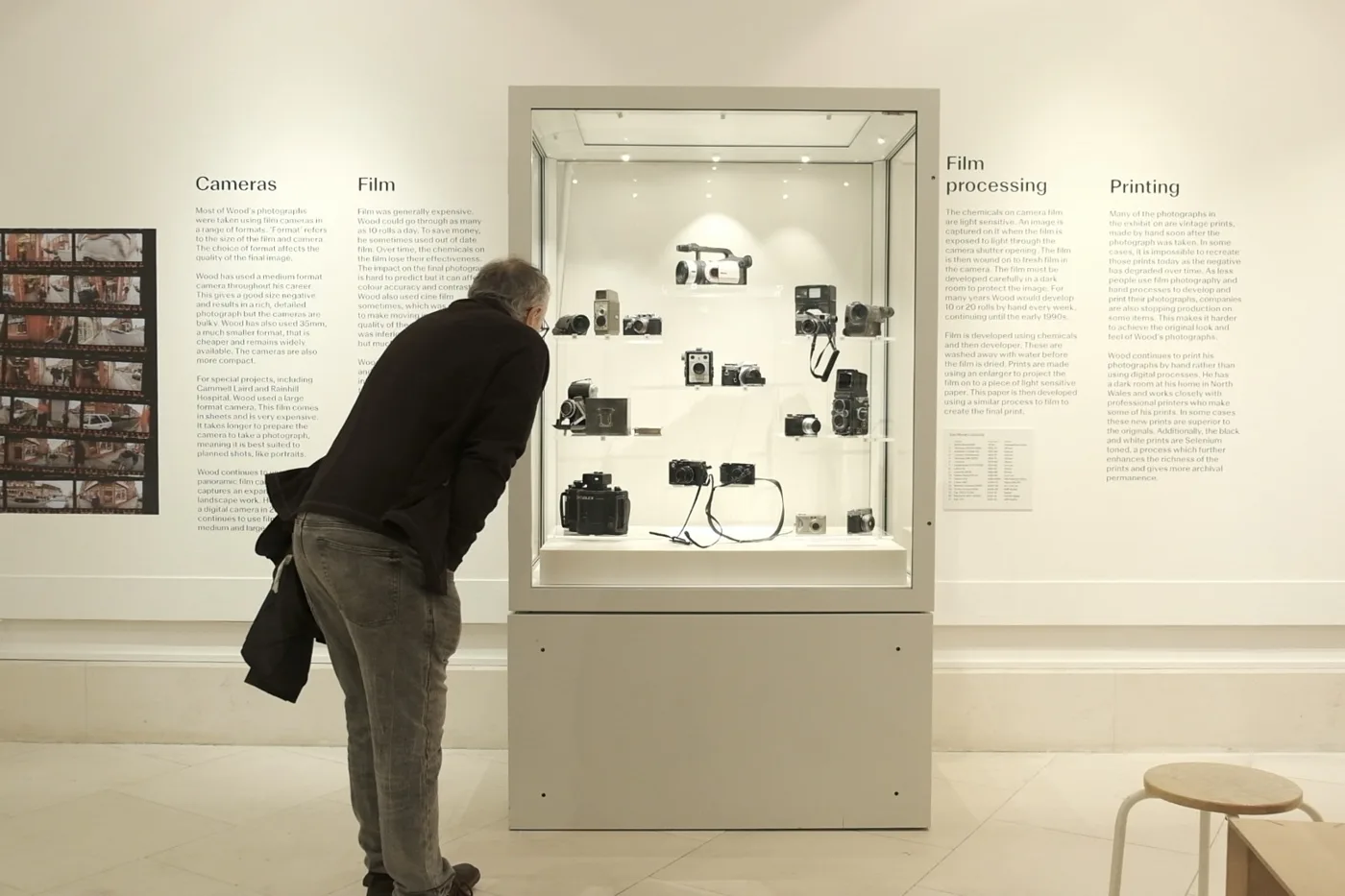 A man in a dark jacket leans in to look at a gallery display case of cameras beside wall panels about cameras, film, processing and printing