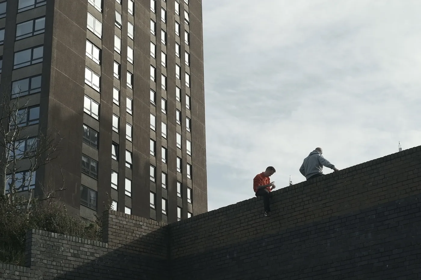 Two people sit on top of a high brick wall beside a tower block under a grey sky