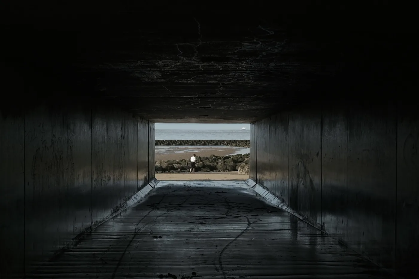 Looking out of a dark concrete underpass towards the sea and a sea wall in daylight