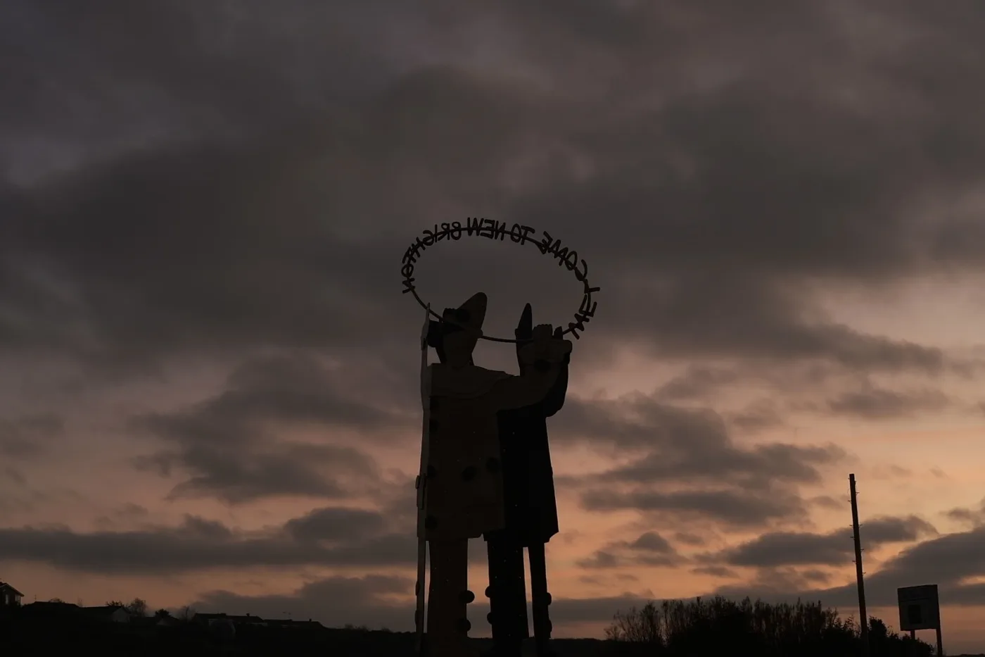 A silhouetted sculpture of a figure with a ring of metal lettering above its head against an orange dusk sky